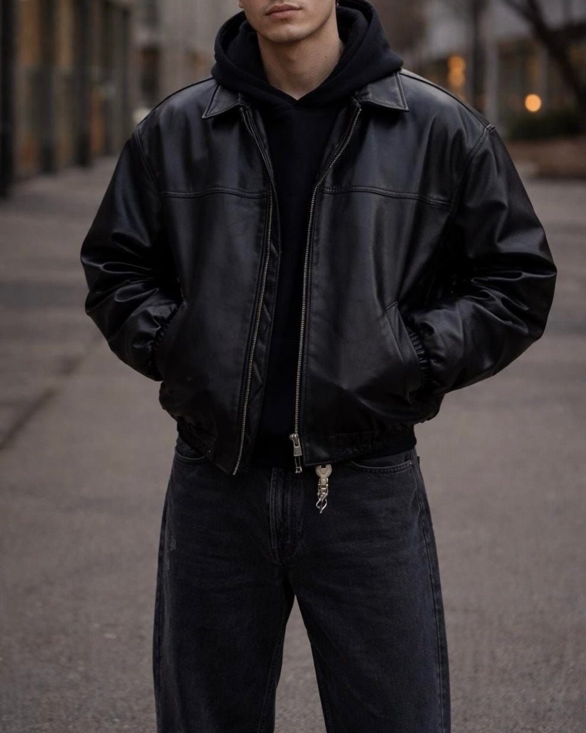 A person wearing a black leather jacket over a black hoodie and black jeans outdoors.