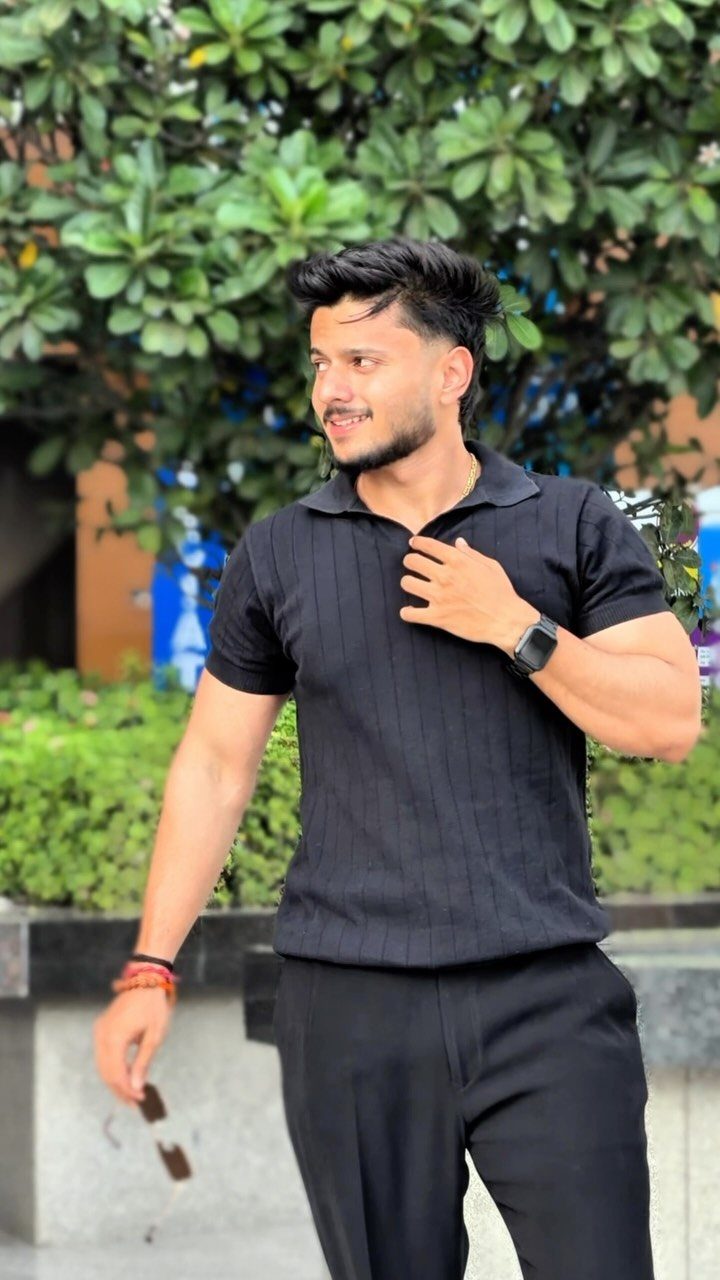 A man in a black polo shirt and black trousers posing against green trees.