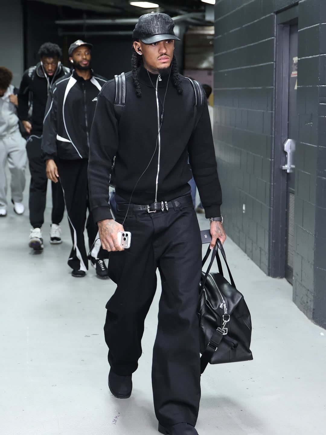 A man walking in a hallway wearing an all-black jacket, wide trousers, and a cap.