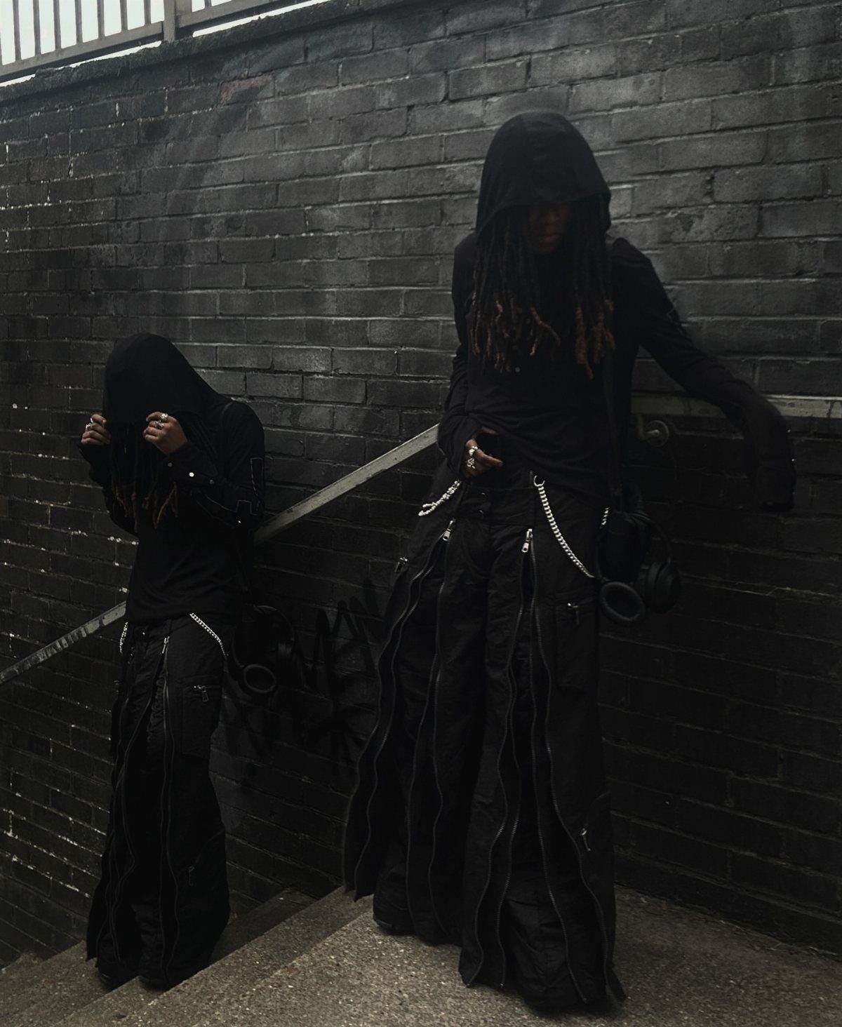 Two people wearing all-black hooded outfits and wide-leg trousers standing on outdoor stairs.