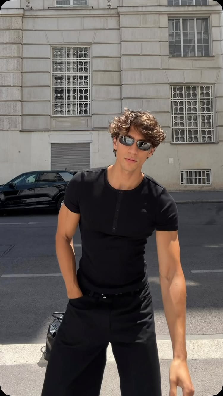 A young man posing in a tight black t-shirt, black pants, and sunglasses on a city street.