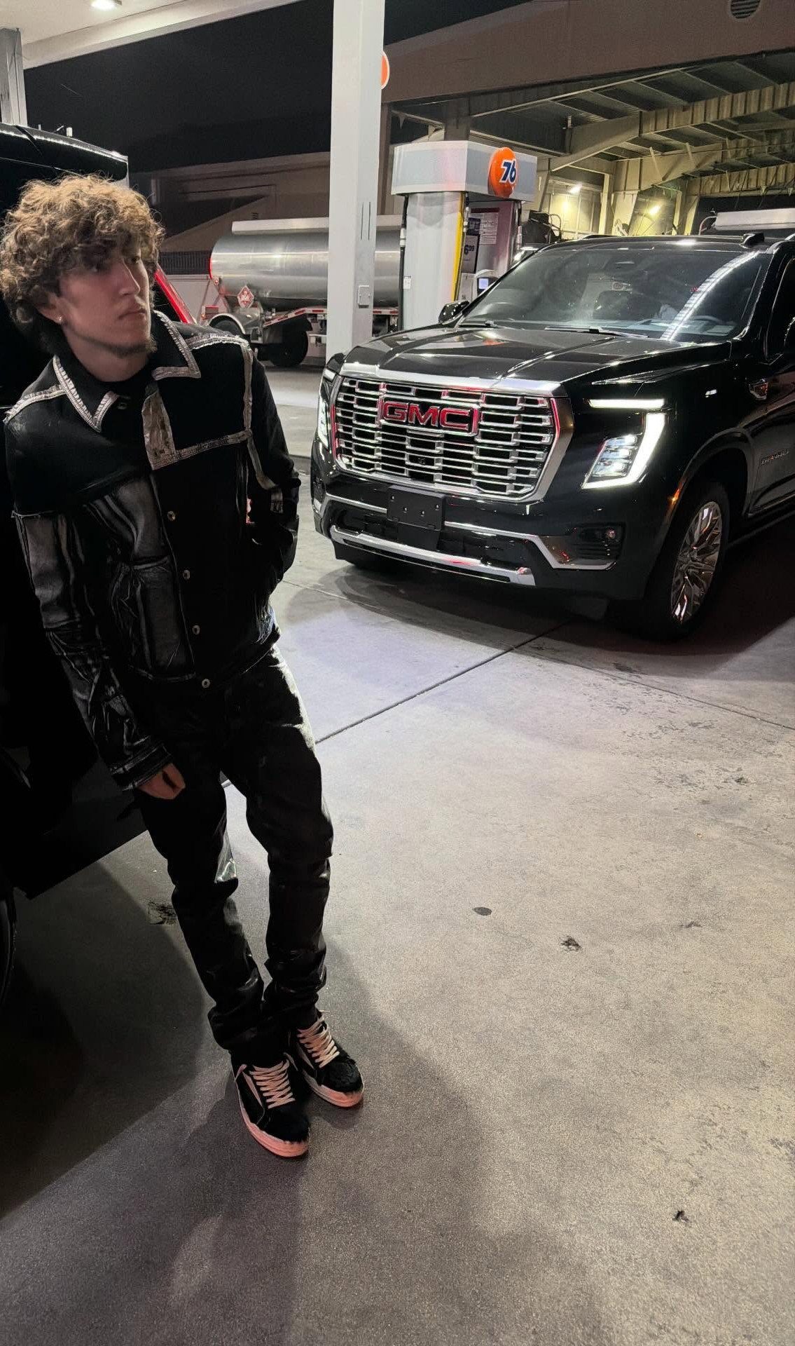 A young man in a black leather jacket and black pants leans against a car at night.