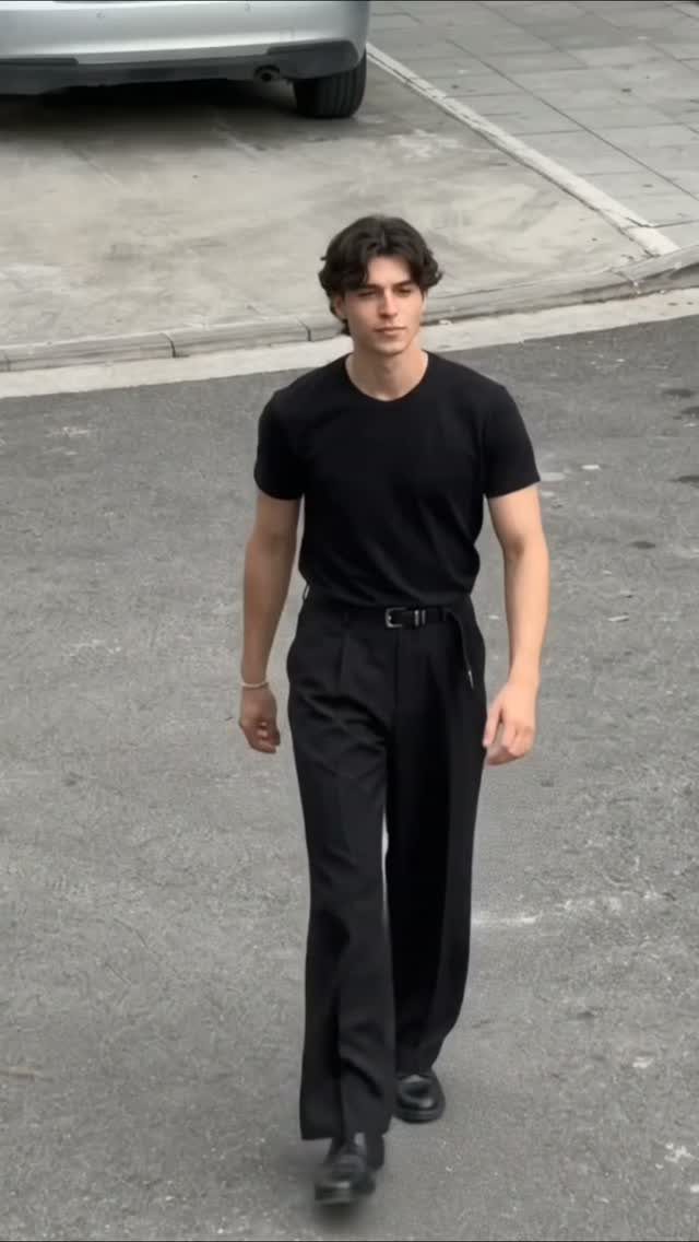 A young man walking outdoors wearing an all-black outfit consisting of a t-shirt and wide-leg trousers.