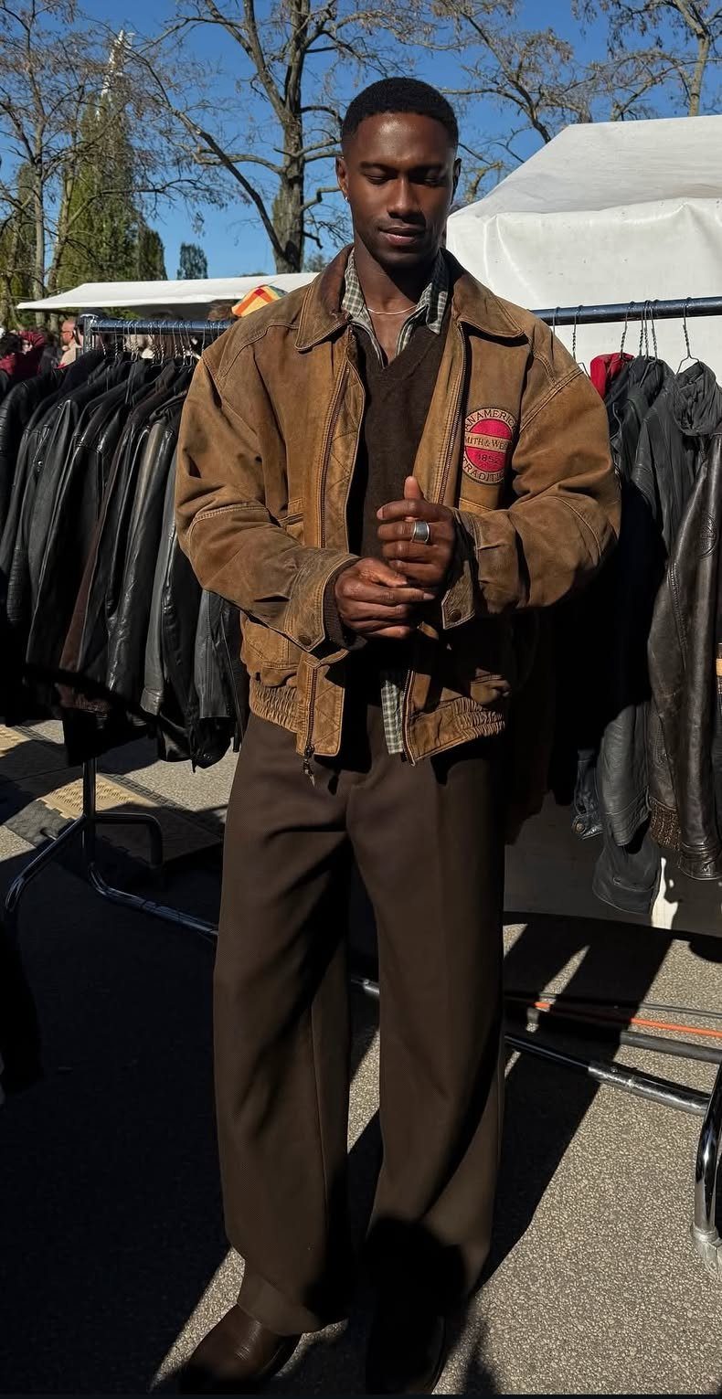 A man wearing a brown suede jacket, brown sweater, and brown trousers standing outdoors.
