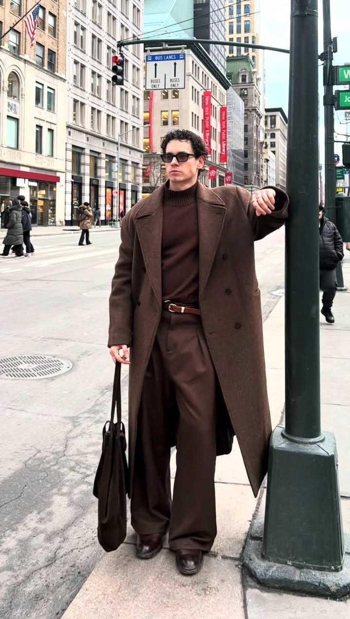 Man in a long brown coat, brown turtleneck, and brown trousers standing on a city street.