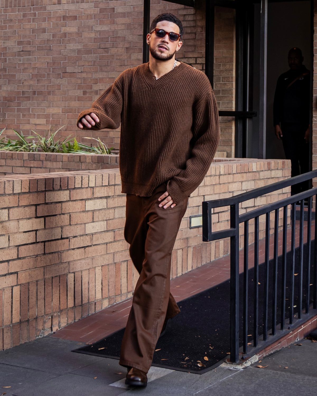 Man in a brown ribbed sweater and brown trousers walking outdoors near a brick building.