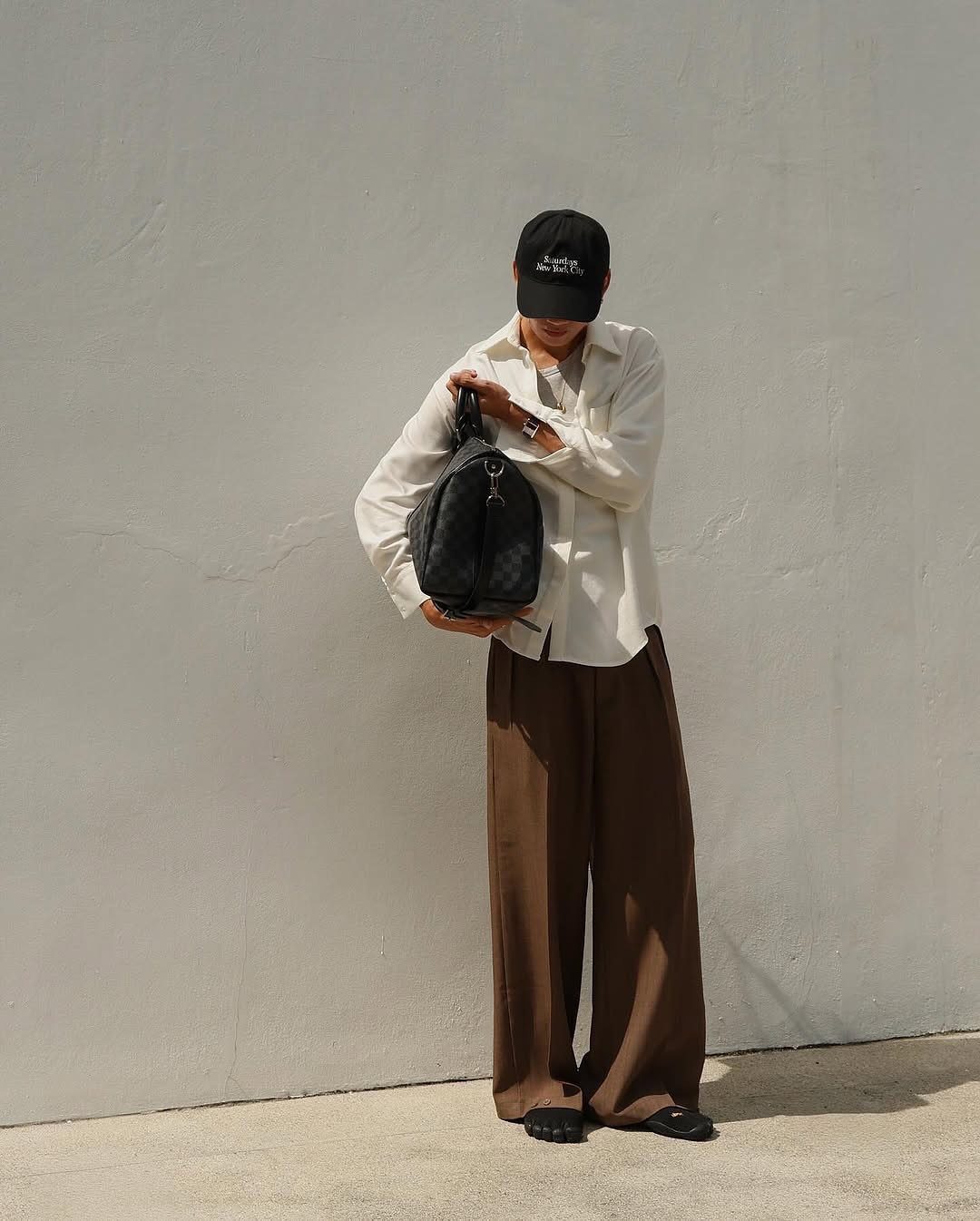 A person wearing an off-white shirt and wide-leg brown trousers standing against a white wall.