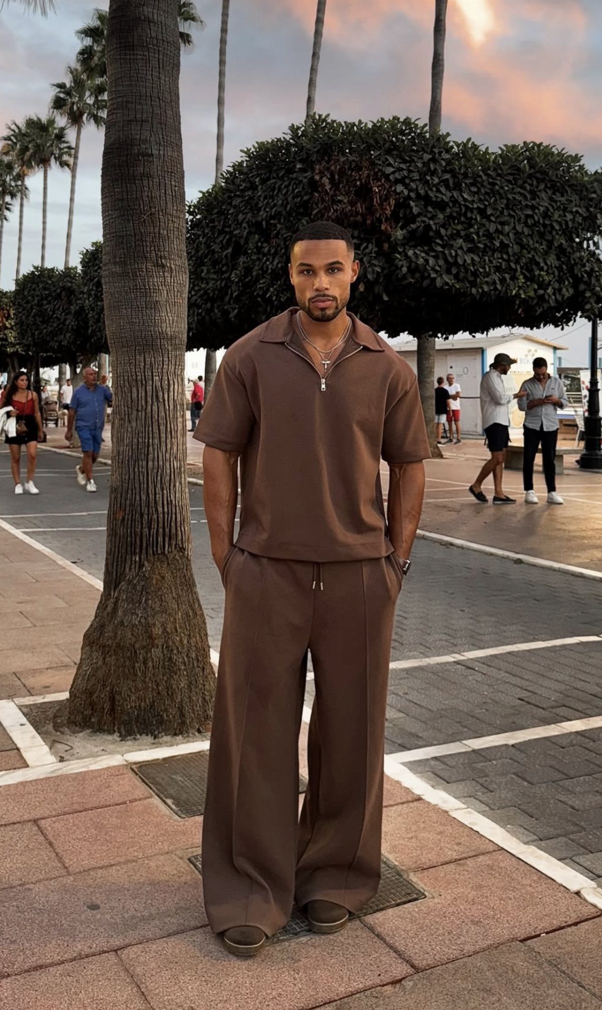 A man wearing a matching brown short-sleeved polo and wide-leg trousers standing by palm trees.