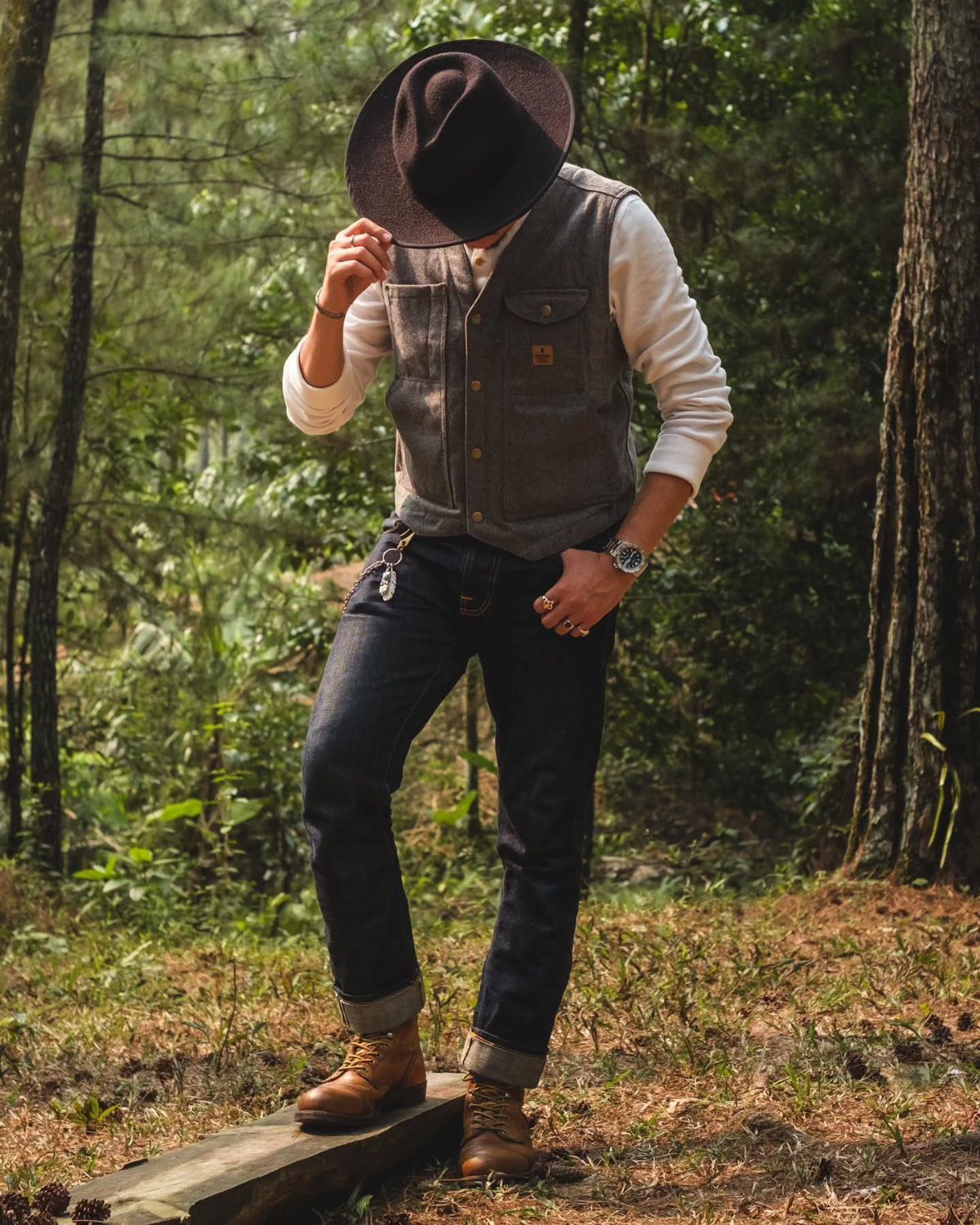 A person wearing a grey vest, white shirt, dark jeans, and a wide-brimmed hat in a forest.