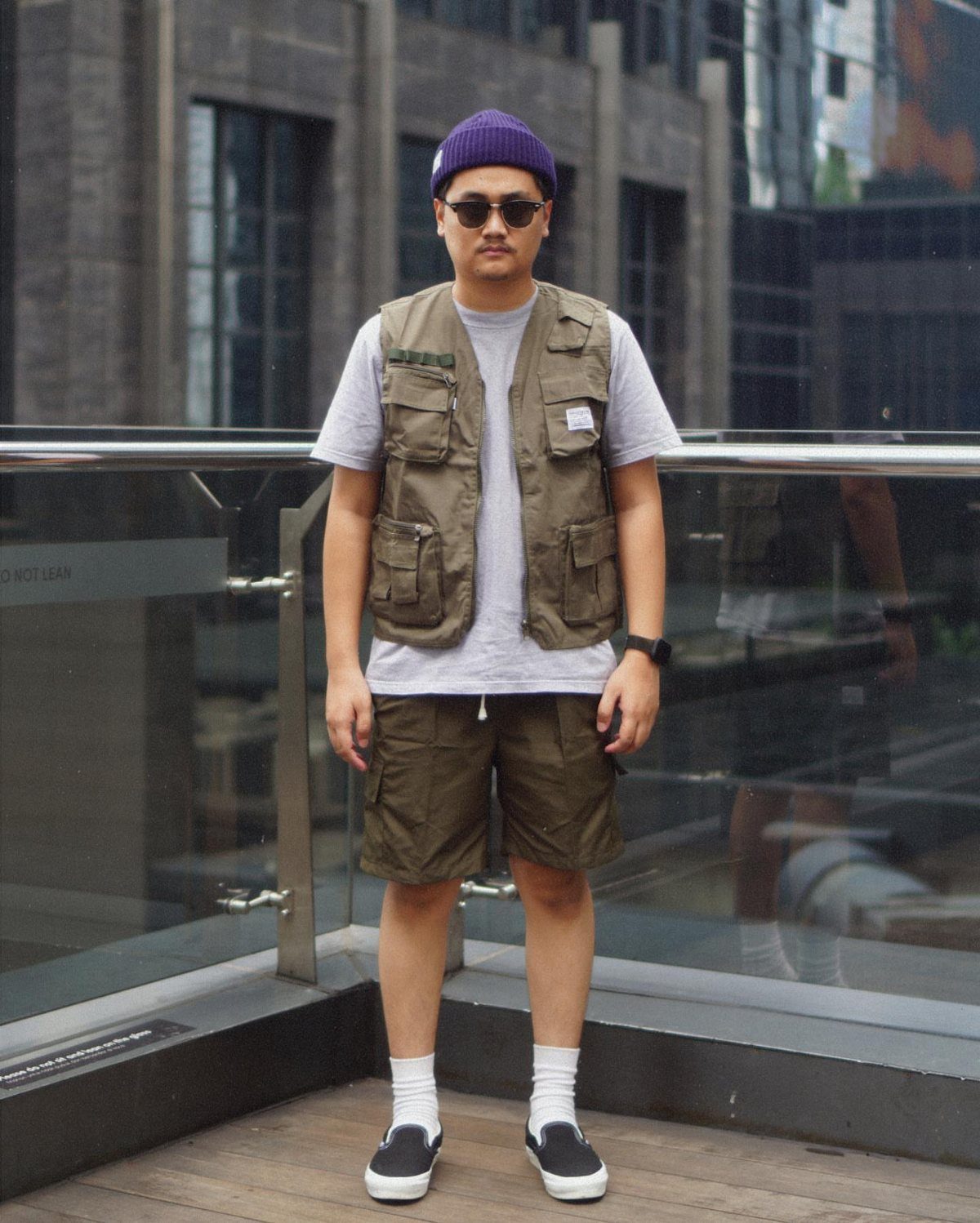 A man standing outdoors wearing a grey t-shirt, olive green utility vest, and matching shorts.