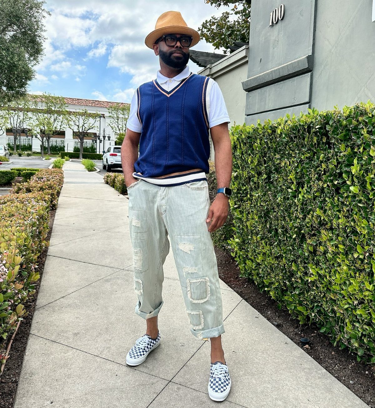 Man in a navy knit vest and distressed light denim jeans standing on a sidewalk.