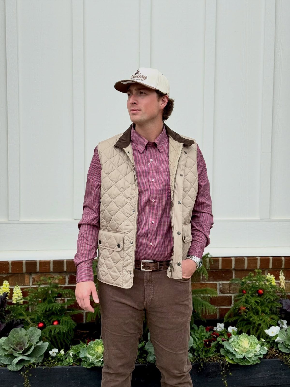 A man wearing a cream quilted vest, maroon shirt, and brown trousers standing outdoors.