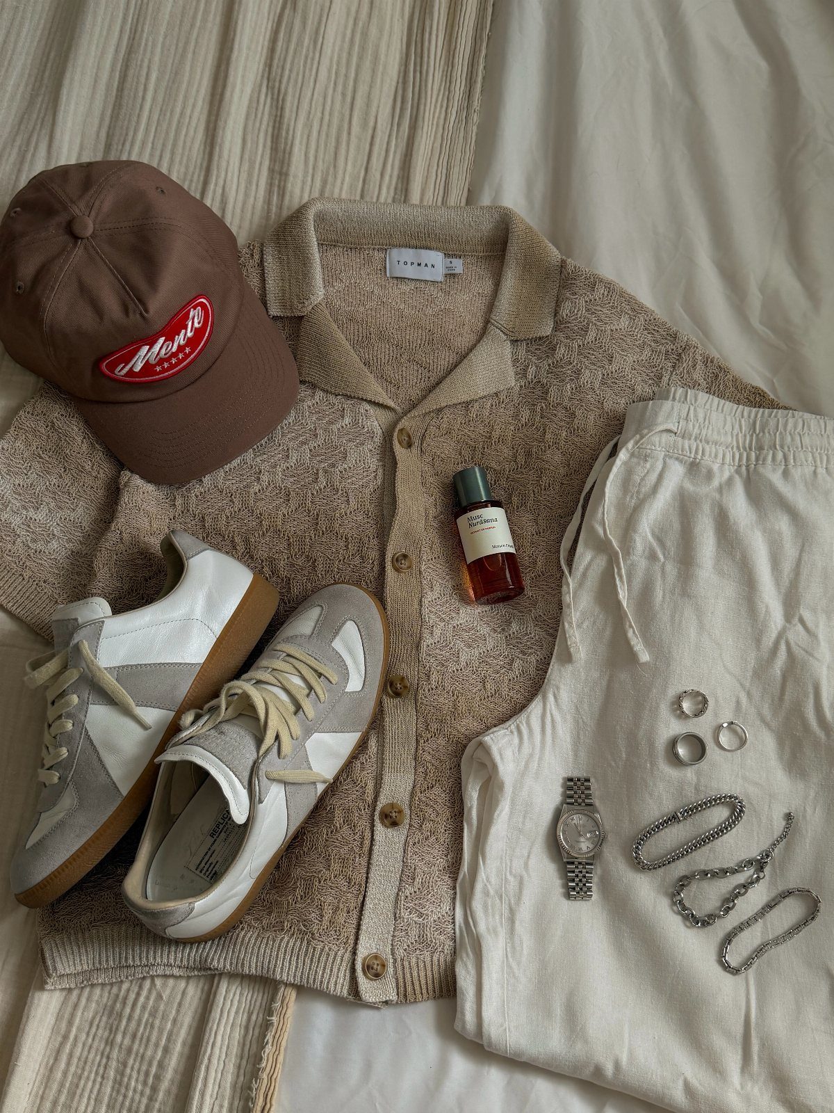Men's clothing flat lay featuring a beige cardigan, brown cap, white shorts, sneakers, and silver accessories.