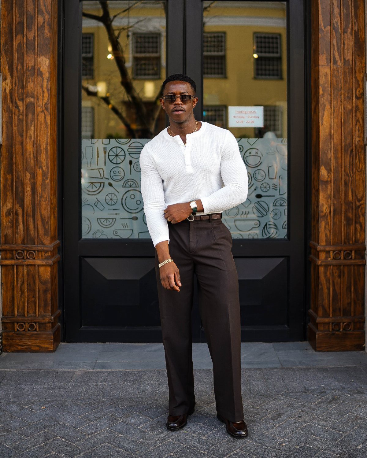 Man in a white textured shirt and brown trousers standing before dark doors.