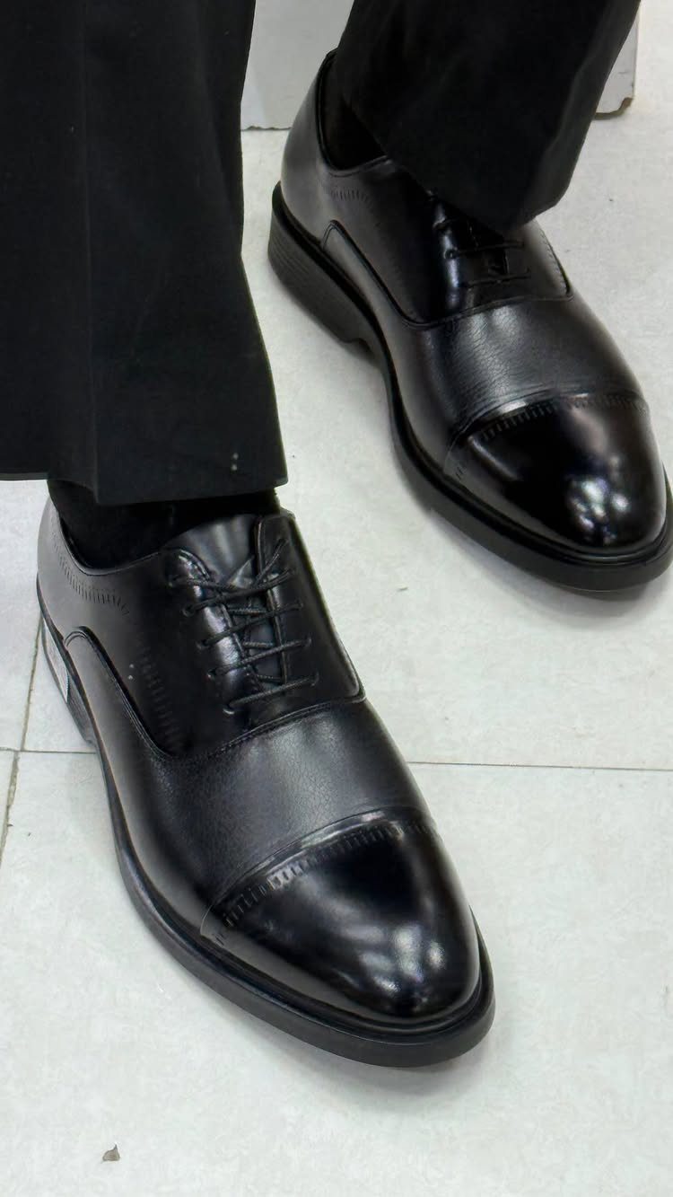 Black leather derby dress shoes worn with black trousers on a tiled floor.