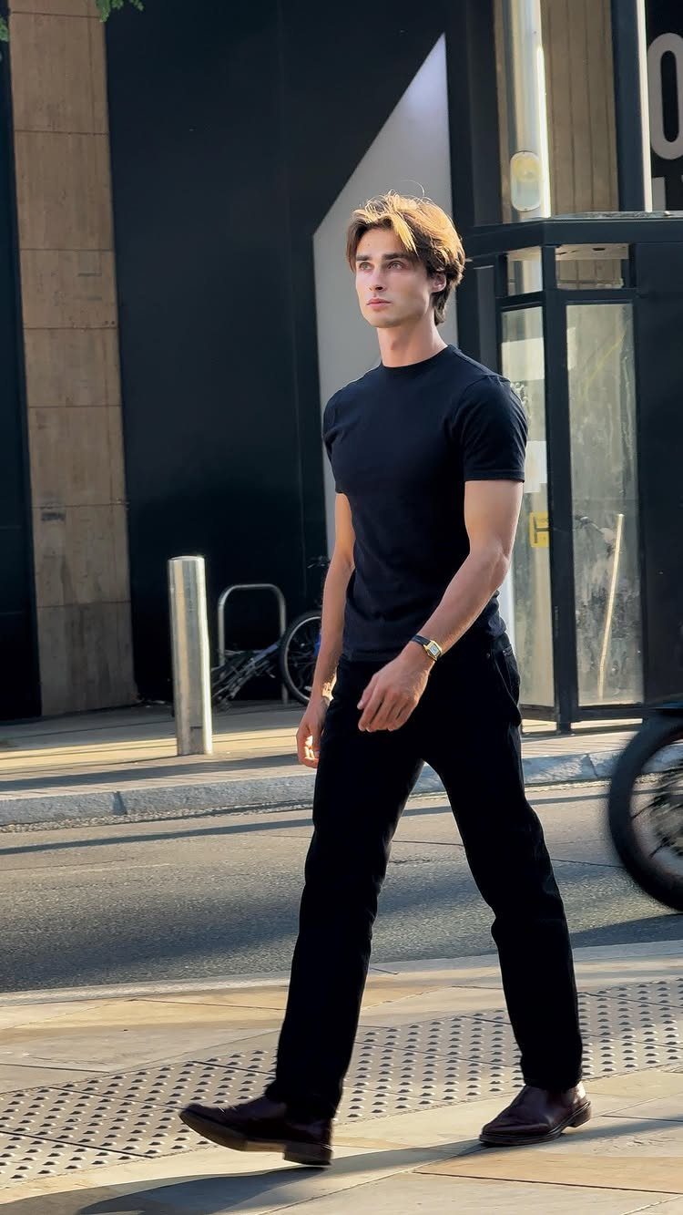 A young man walking outdoors wearing a black t-shirt, black pants, and dark leather shoes.