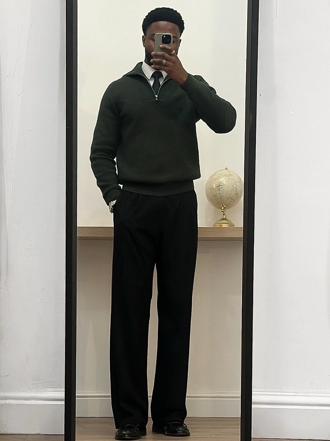 Man in dark green sweater, black tie, and wide-leg trousers posing in a mirror.