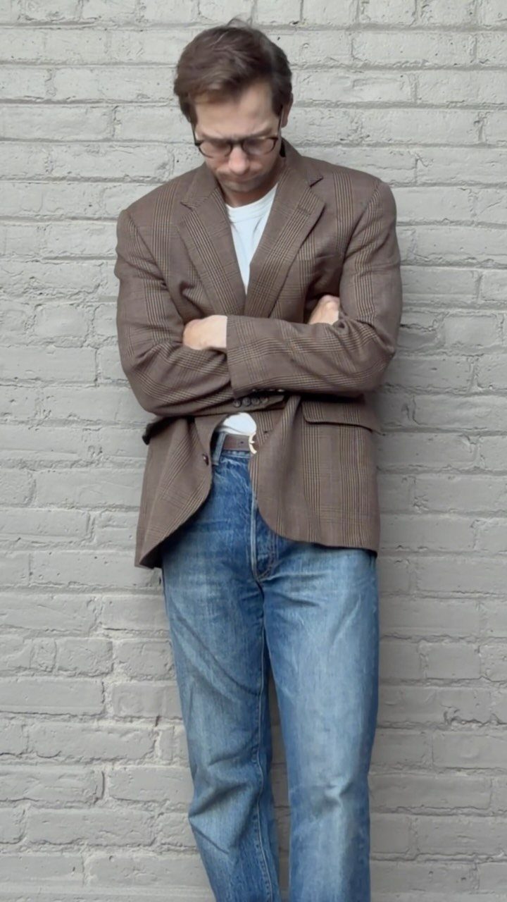 Man wearing a brown patterned sport coat, white t-shirt, and blue jeans against a brick wall.