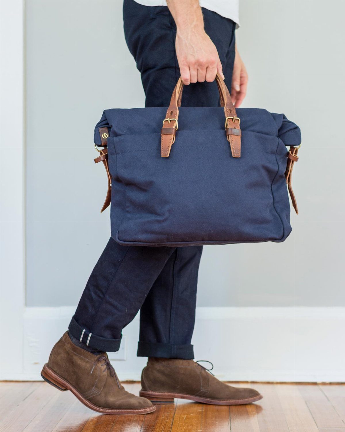 Man wearing navy trousers, brown suede boots, and carrying a large navy canvas bag.
