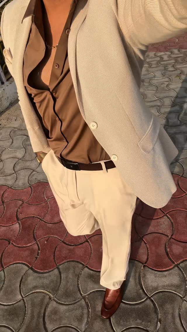 Man wearing a beige blazer, brown shirt, and cream trousers standing on a stone path.