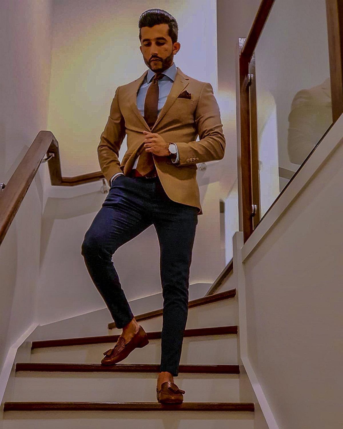 Man in a tan blazer, blue shirt, and navy chinos posing on a staircase.