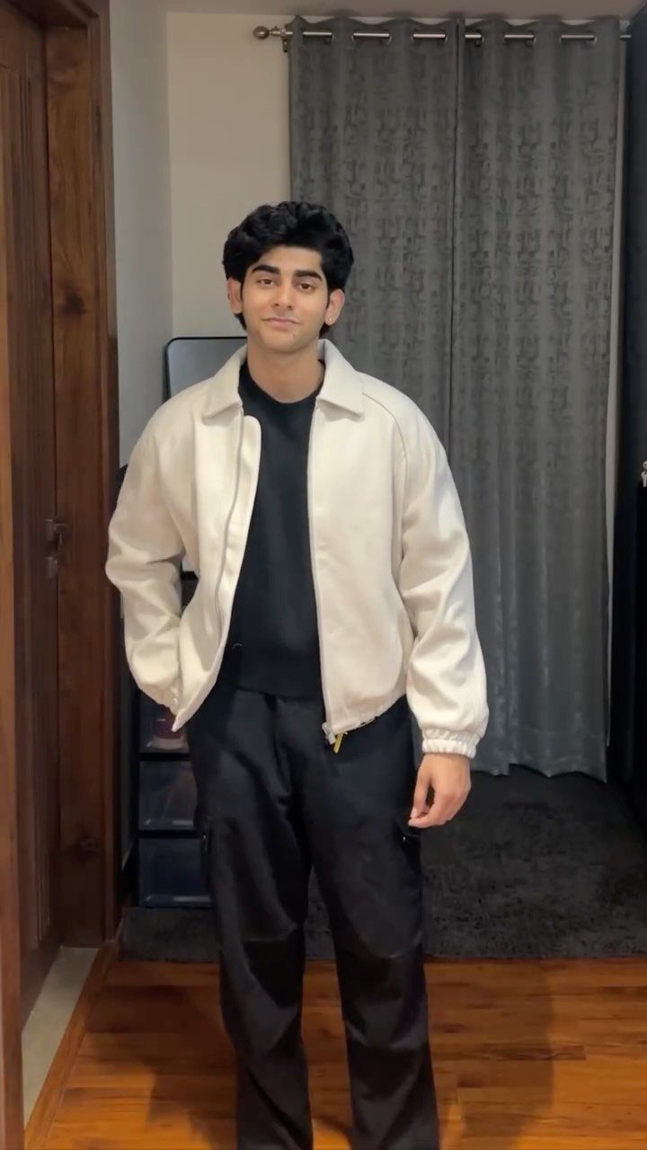 A young man posing in a cream beige jacket, black t-shirt, and black trousers.