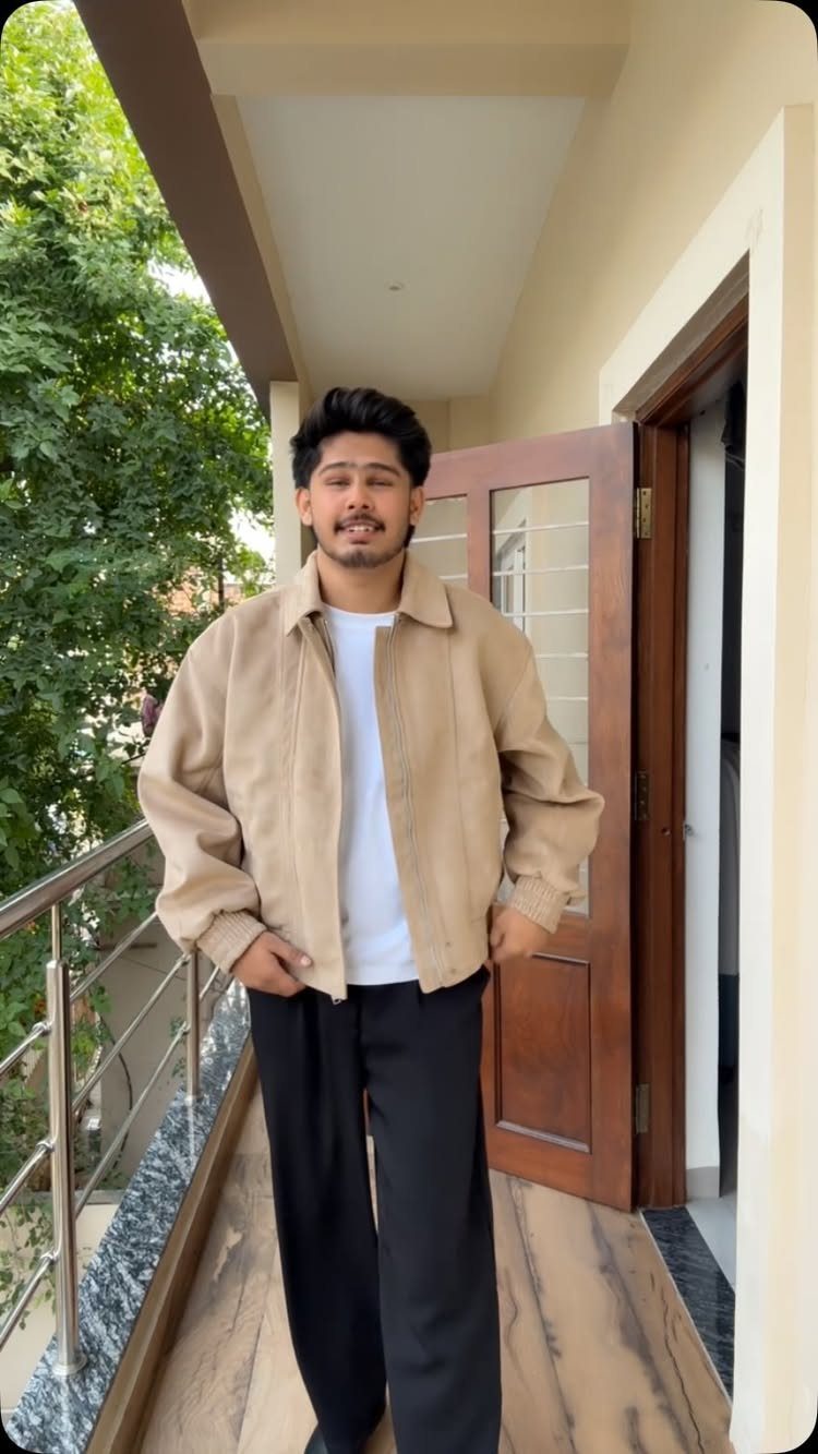 A smiling man wearing a beige jacket, white t-shirt, and black trousers standing on a balcony.