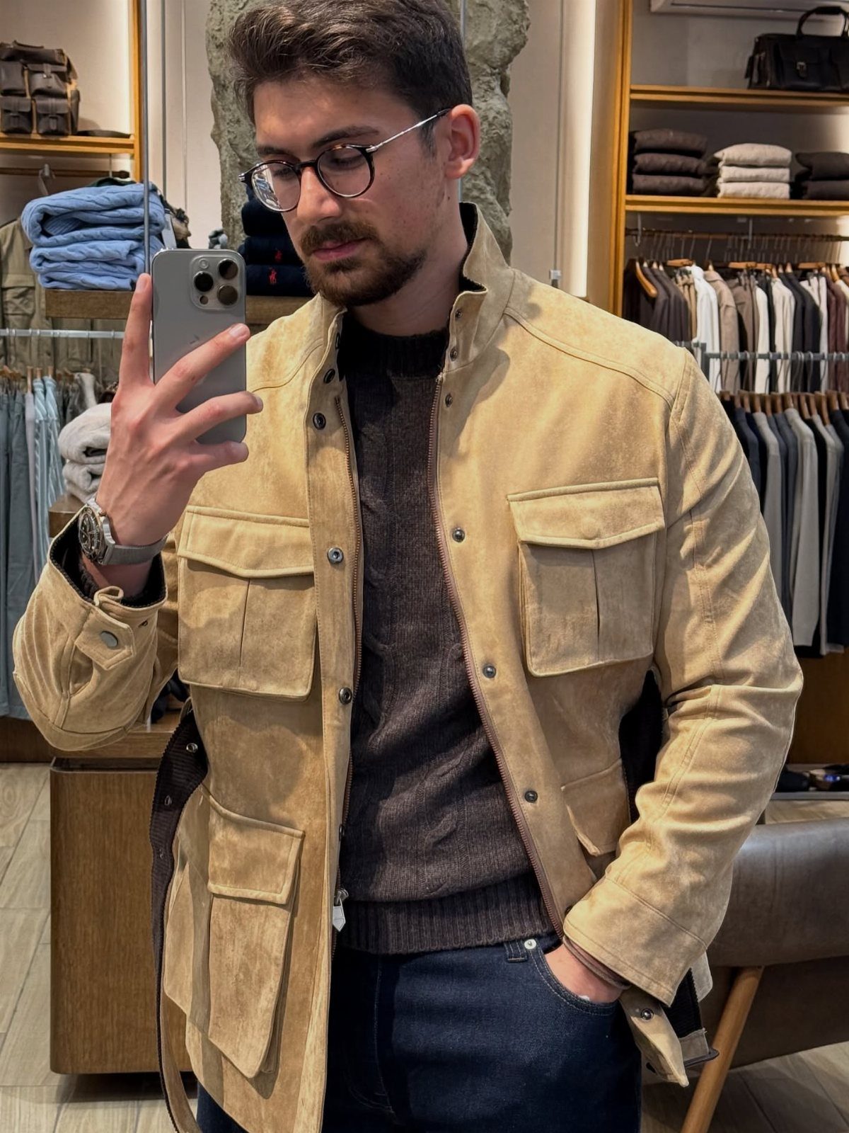 Man in a beige suede jacket, dark sweater, and blue jeans taking a selfie.