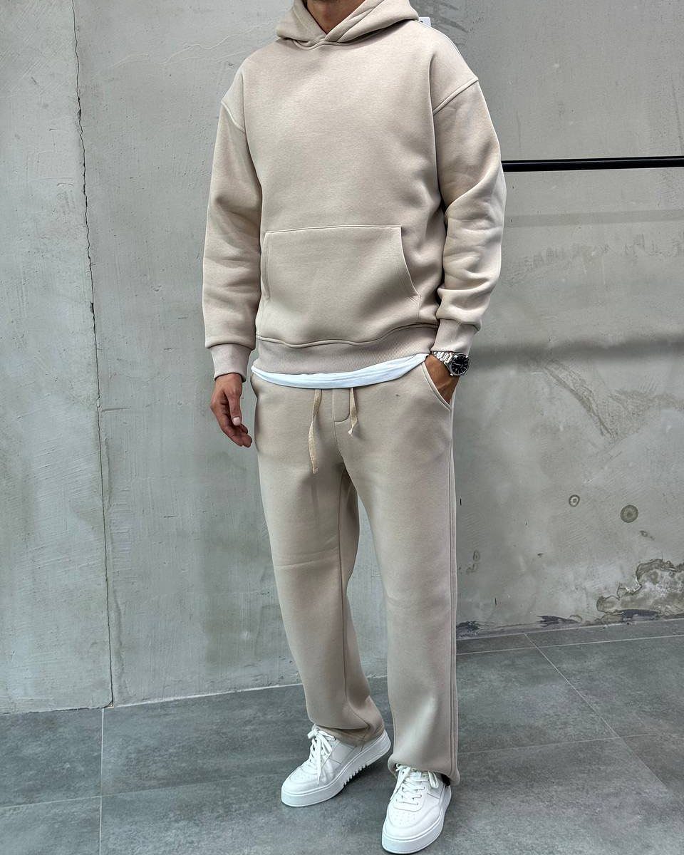 A person wearing a matching beige hoodie and sweatpants set with white sneakers against a concrete wall.