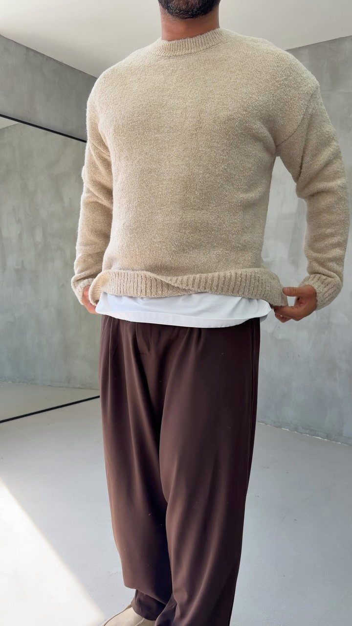 A man wearing a beige textured sweater with brown trousers and a white undershirt.