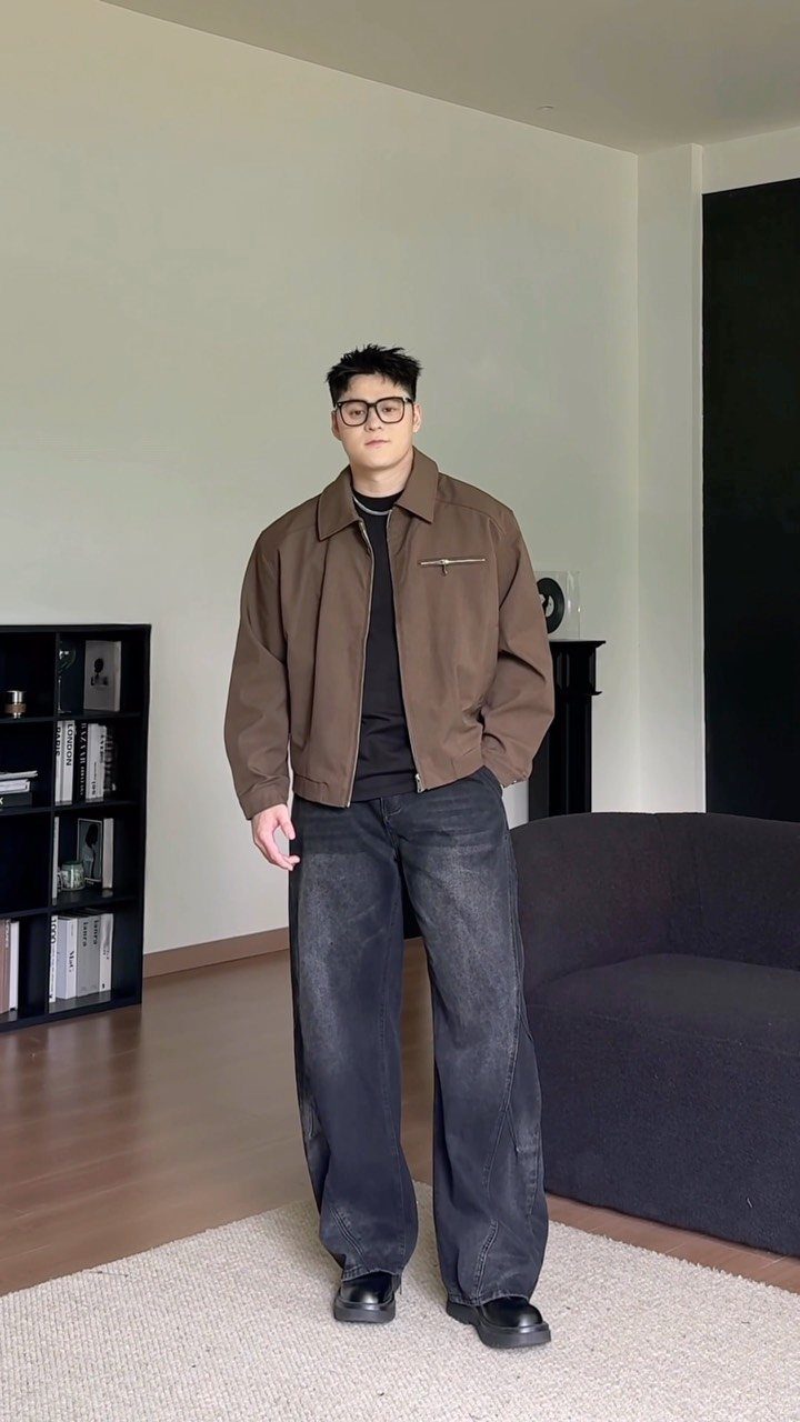 Man wearing a brown jacket and oversized, wide-leg black denim jeans standing indoors.