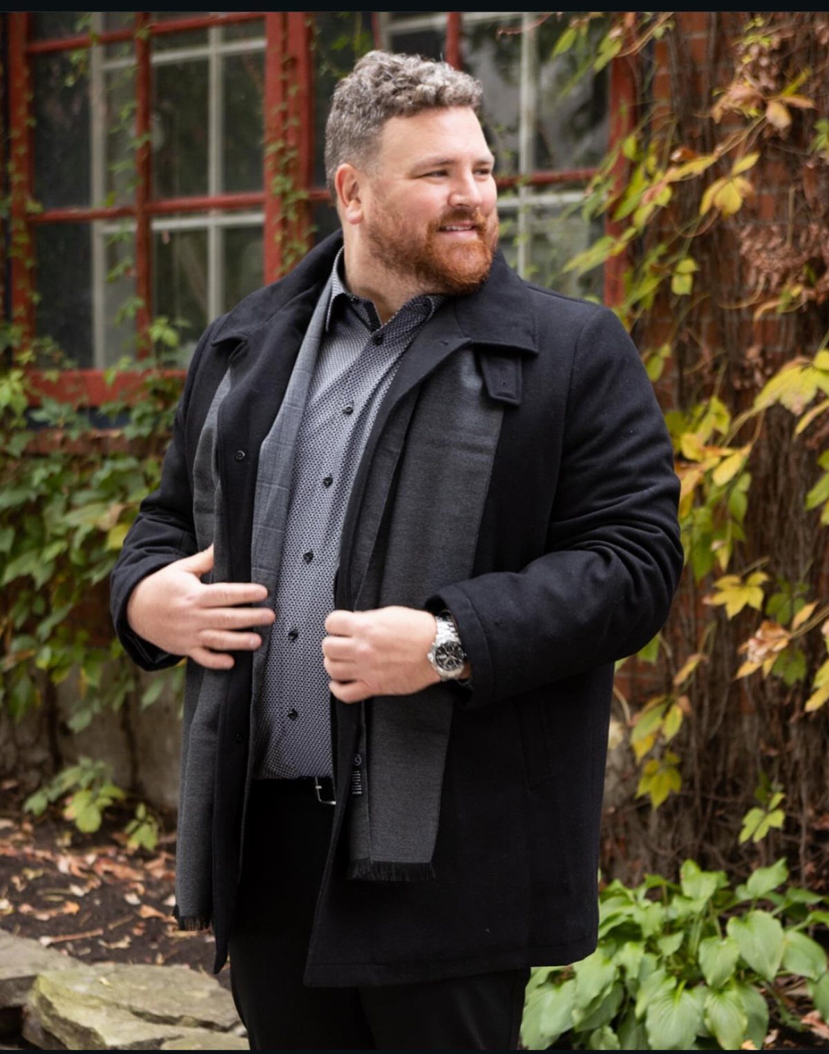 A bearded man wearing a black coat and grey scarf standing outdoors near autumn leaves.