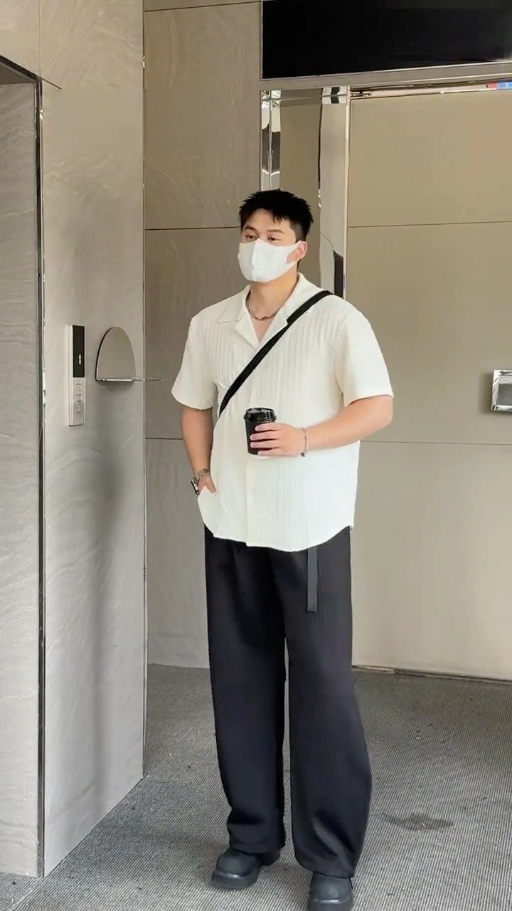 Man in a white short-sleeved shirt and wide-leg black trousers standing in a hallway.