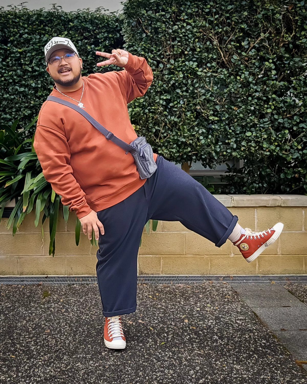 A man in an orange sweatshirt and blue trousers posing playfully in front of a green hedge.
