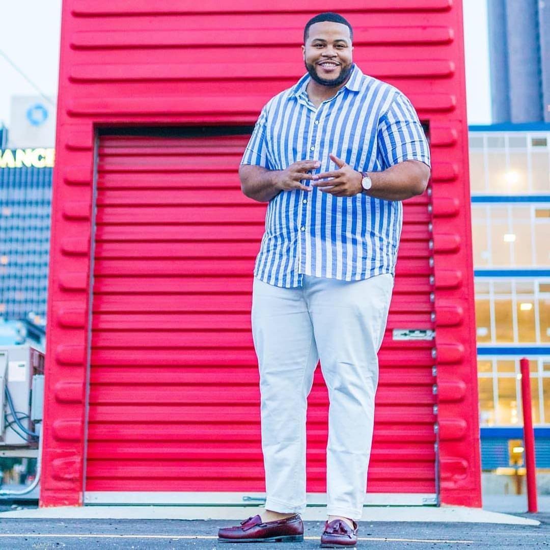 Smiling man in a blue striped shirt and light grey trousers standing before a red wall.