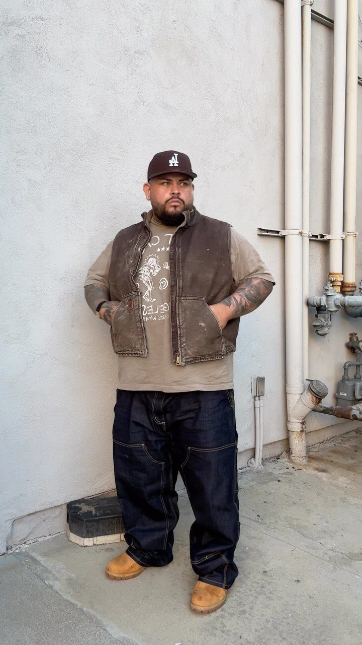 A large man wearing a brown utility vest, tan t-shirt, and dark denim pants standing outdoors.