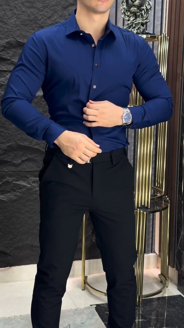Man wearing a navy blue button-down shirt and black trousers standing indoors.