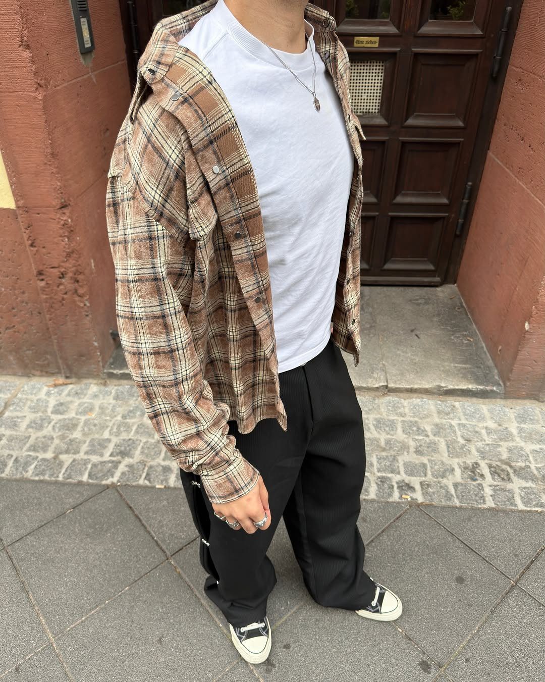 A person wearing a brown and black plaid flannel shirt, white t-shirt, and black trousers.