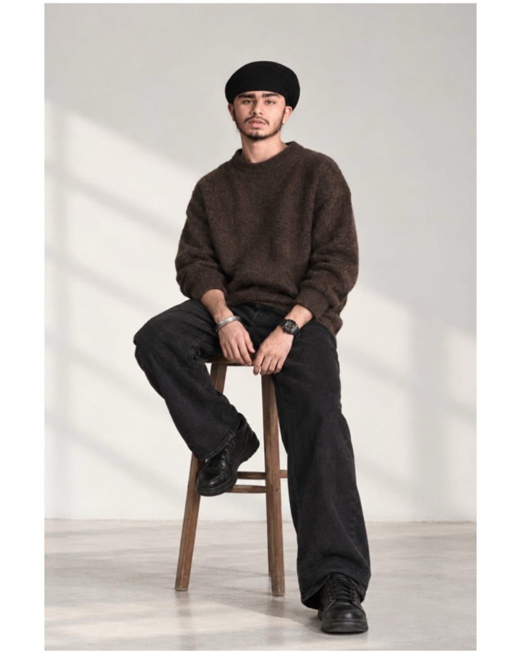 Man in a brown chunky knit sweater and black jeans sitting on a stool.