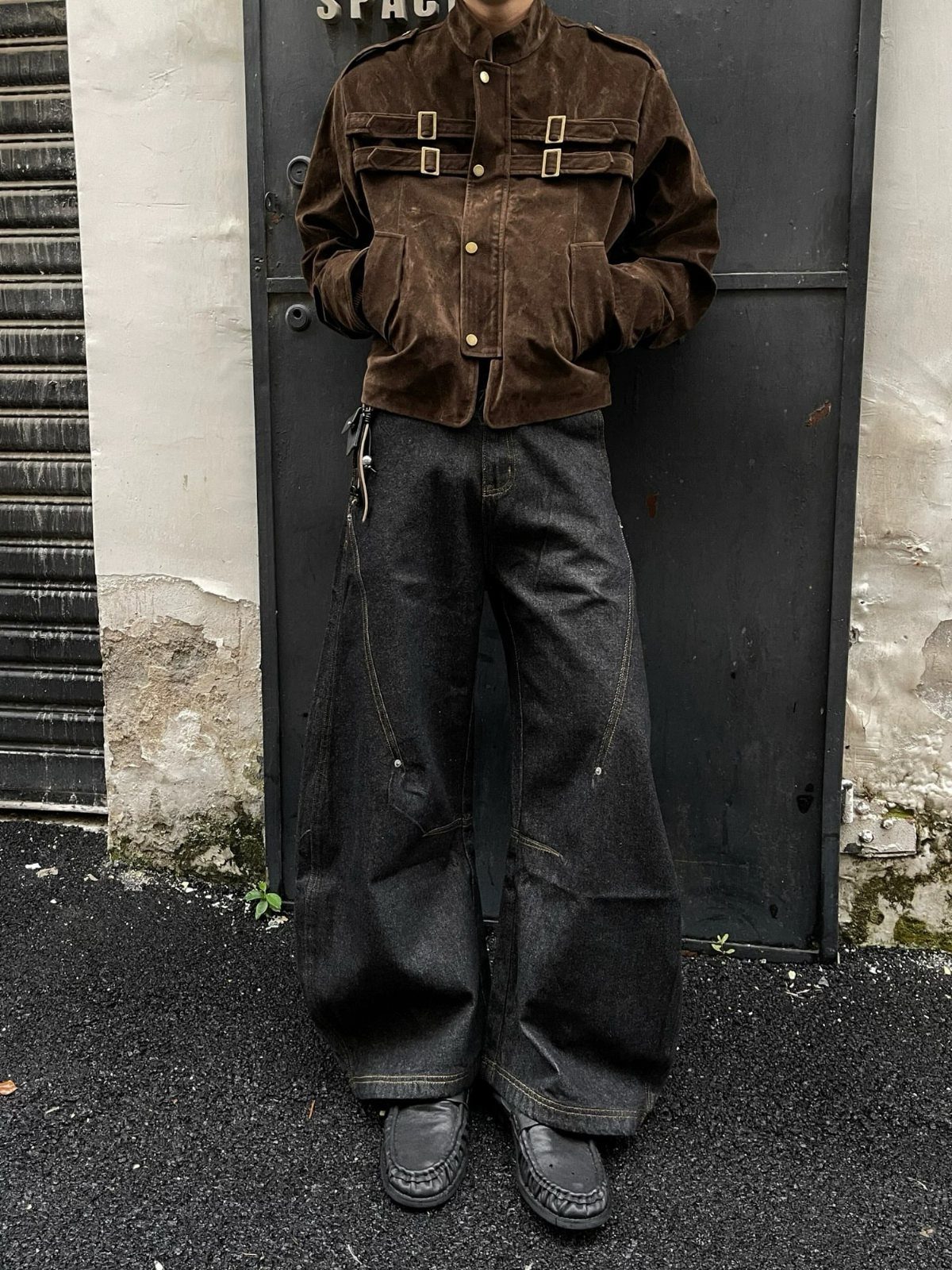 Person wearing a brown suede jacket with buckles and wide black jeans standing outdoors.