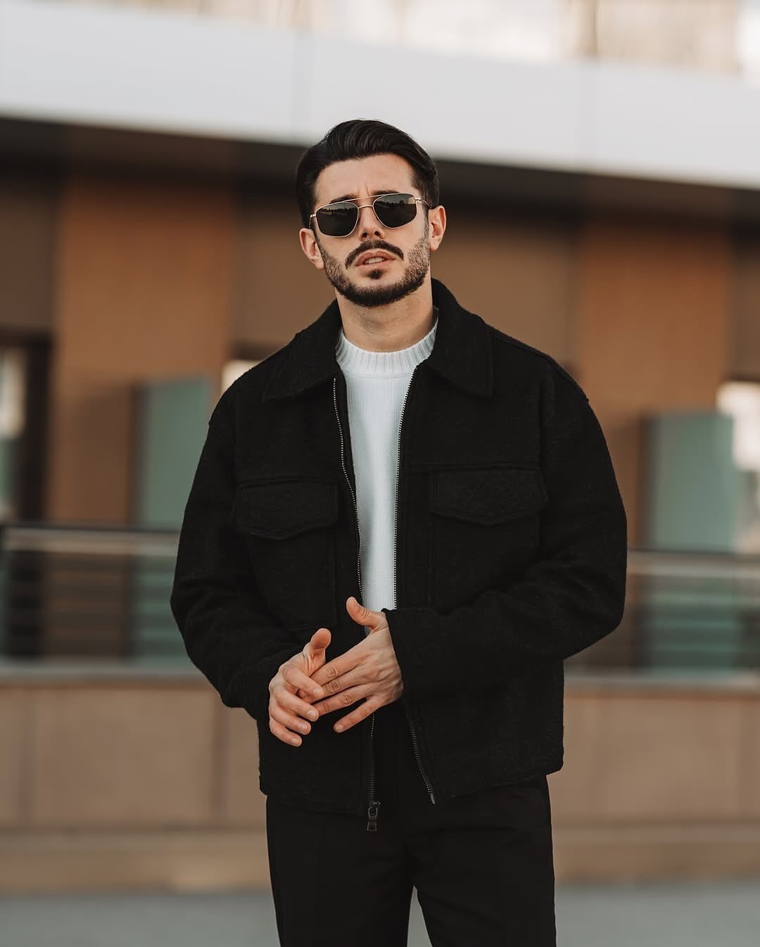Man in a black jacket and white sweater posing outdoors wearing sunglasses.