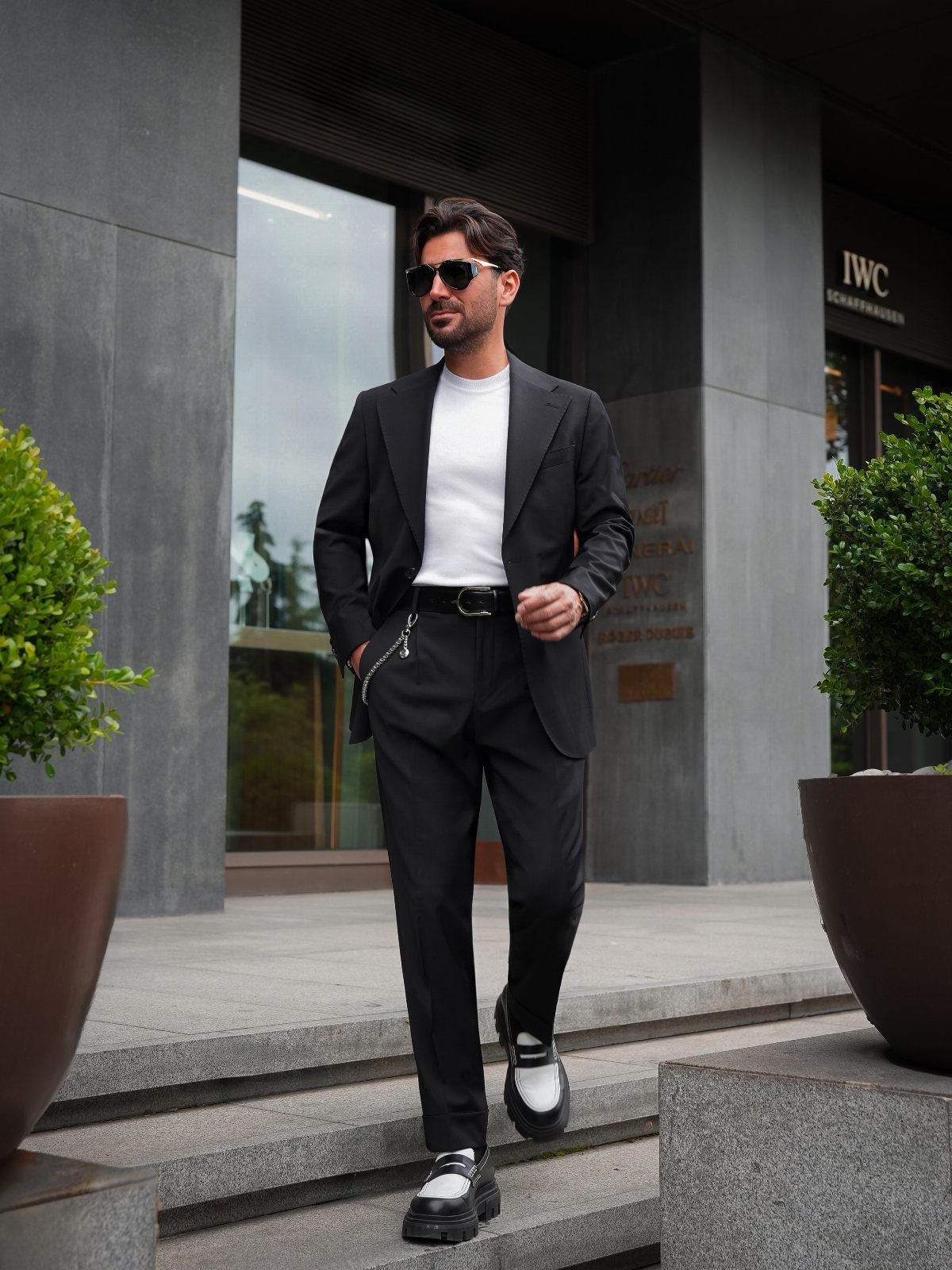 Man in a black blazer and trousers with a white t-shirt walking outdoors.