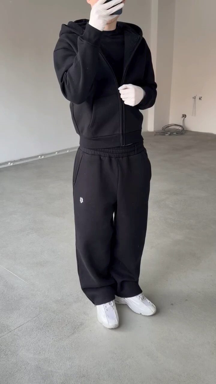 A person dressed in a black hoodie, black joggers, and white sneakers standing in a grey room.