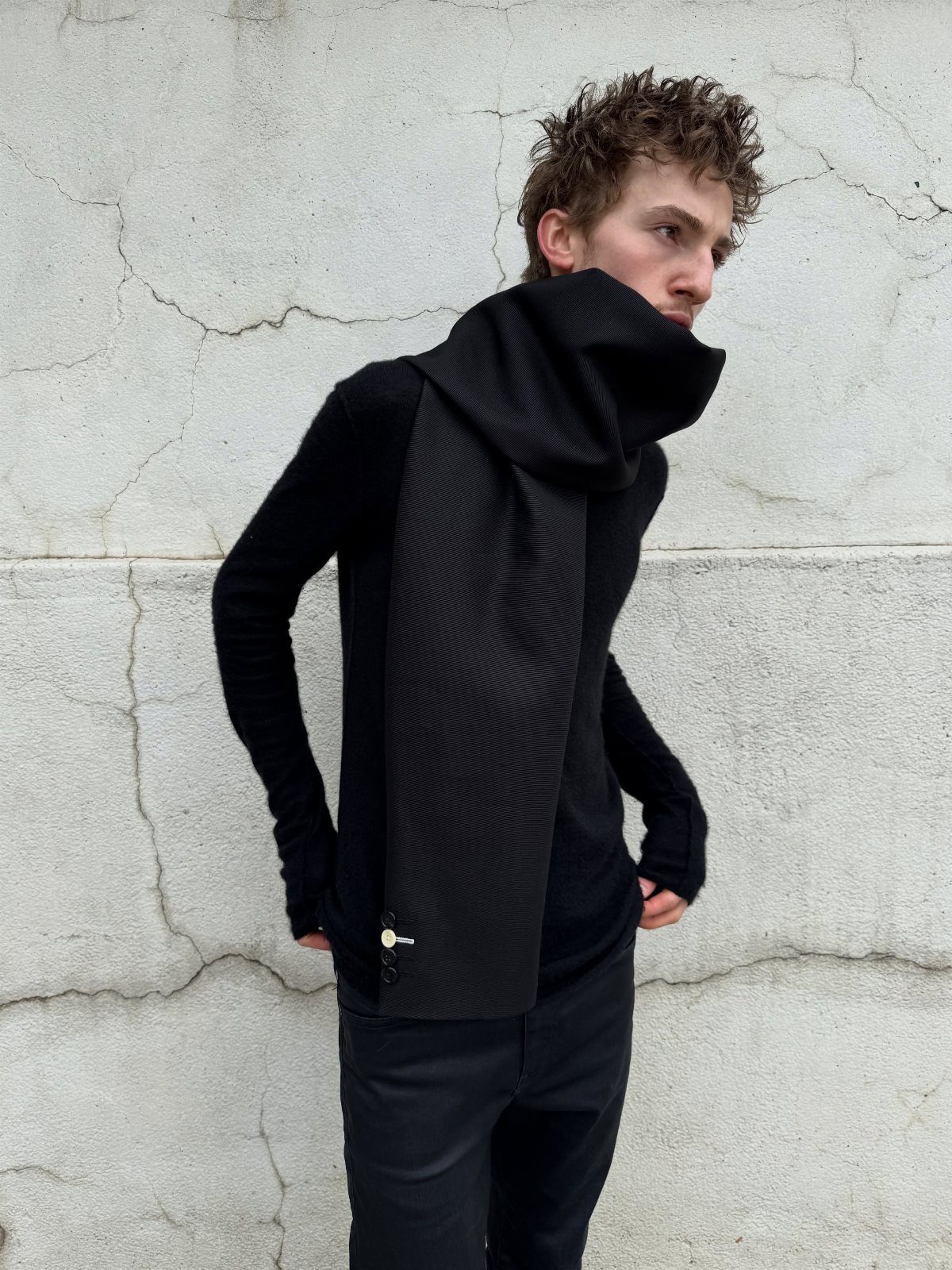 Young man in black knit clothing and a large textured scarf against a concrete wall.