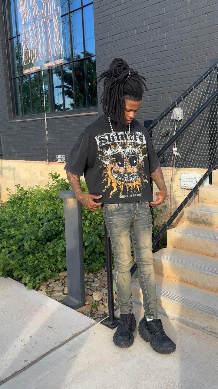 Man wearing a black graphic t-shirt, faded grey jeans, and black sneakers standing outdoors.