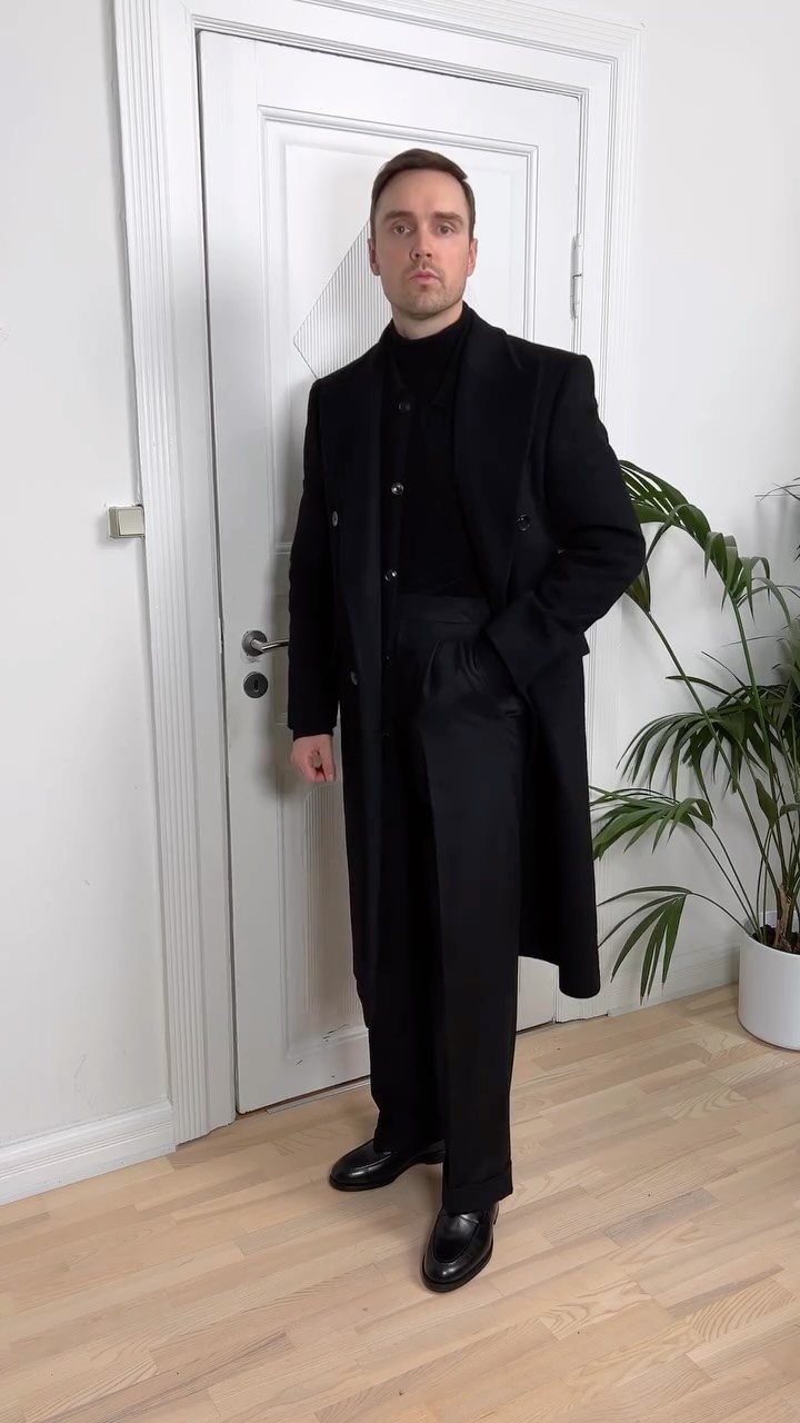 A man posing in an all-black outfit including a long coat and trousers.