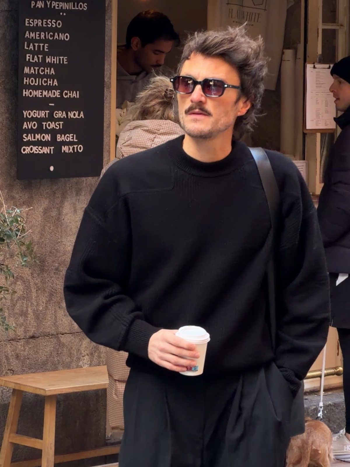 A man in an oversized black sweater and sunglasses holding a coffee cup outdoors.