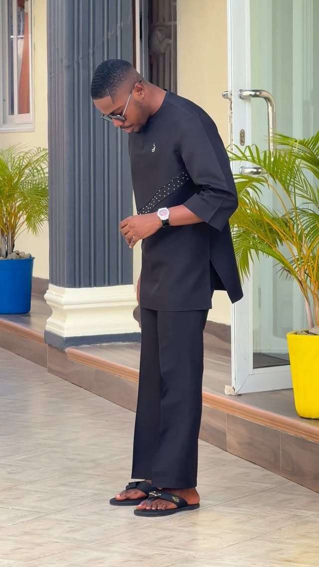 Man in a black tunic and trousers standing near a building with potted plants.