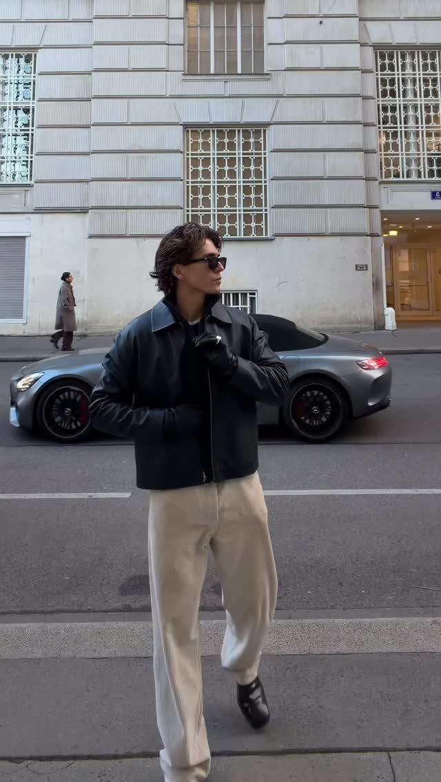 A man posing in a black leather jacket, beige trousers, and sunglasses on a city street.