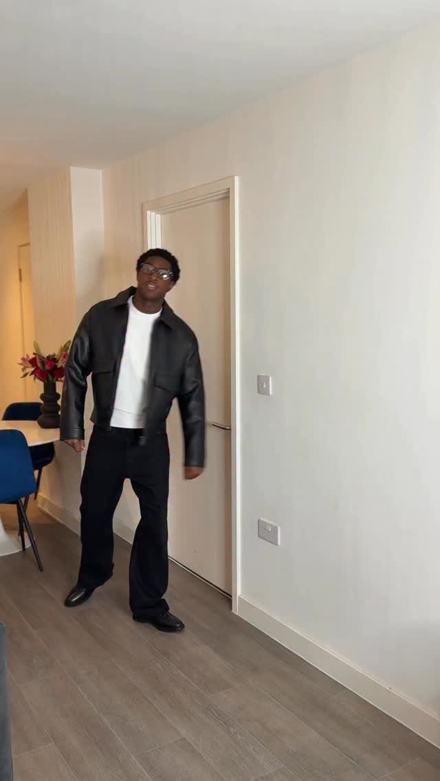 A man wearing a black leather jacket, white t-shirt, and black trousers standing indoors.